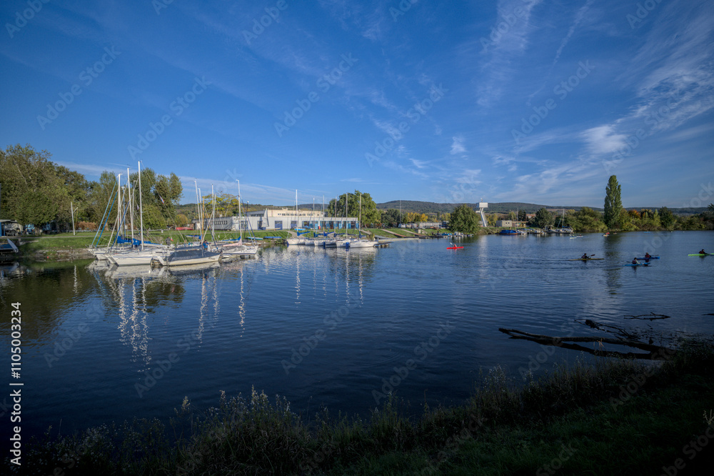 Small marina with sailboats docked on a calm river surrounded by greenery and a clear blue sky. Small marina with sailboats docked on a calm river surrounded by greenery and a clear blue sky.