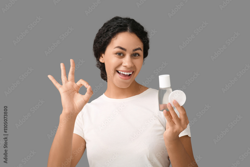 Beautiful young African-American woman with bottle of micellar water and cotton pad showing ok gesture on grey background Beautiful young African-American woman with bottle of micellar water and cotton pad showing ok gesture on grey background