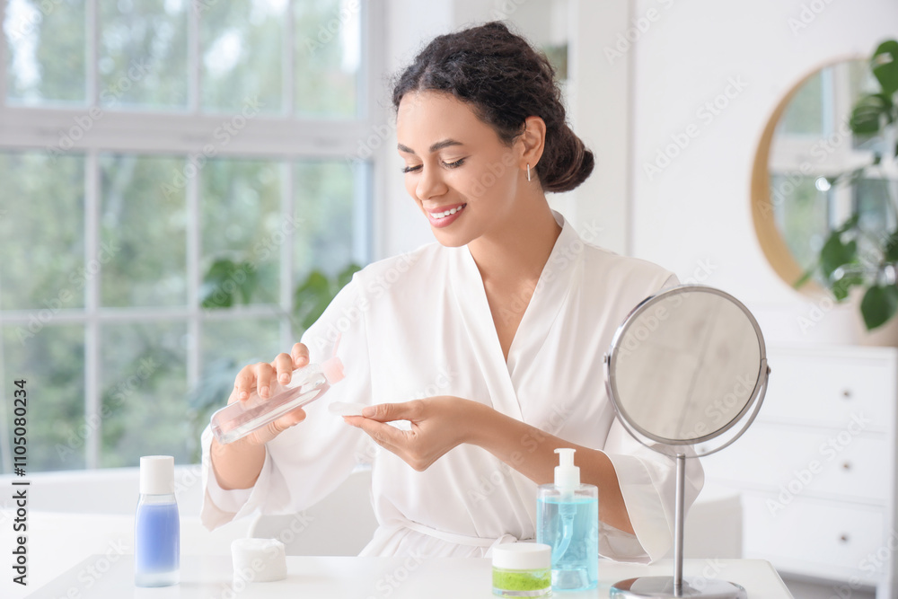 Beautiful young African-American woman applying micellar water on cotton pad at table in bathroom Beautiful young African-American woman applying micellar water on cotton pad at table in bathroom