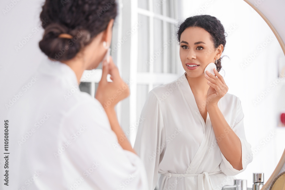 Beautiful young African-American woman removing makeup with cotton pad and looking at mirror in bathroom Beautiful young African-American woman removing makeup with cotton pad and looking at mirror in bathroom