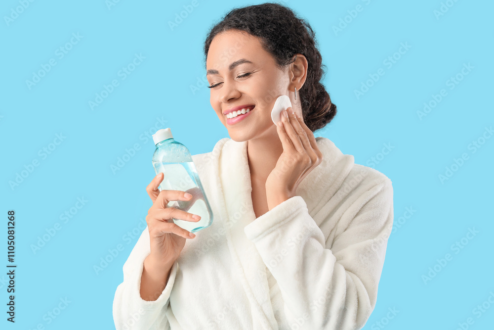 Beautiful young African-American woman removing makeup with cotton pad and holding bottle of micellar water on blue background Beautiful young African-American woman removing makeup with cotton pad and holding bottle of micellar water on blue background
