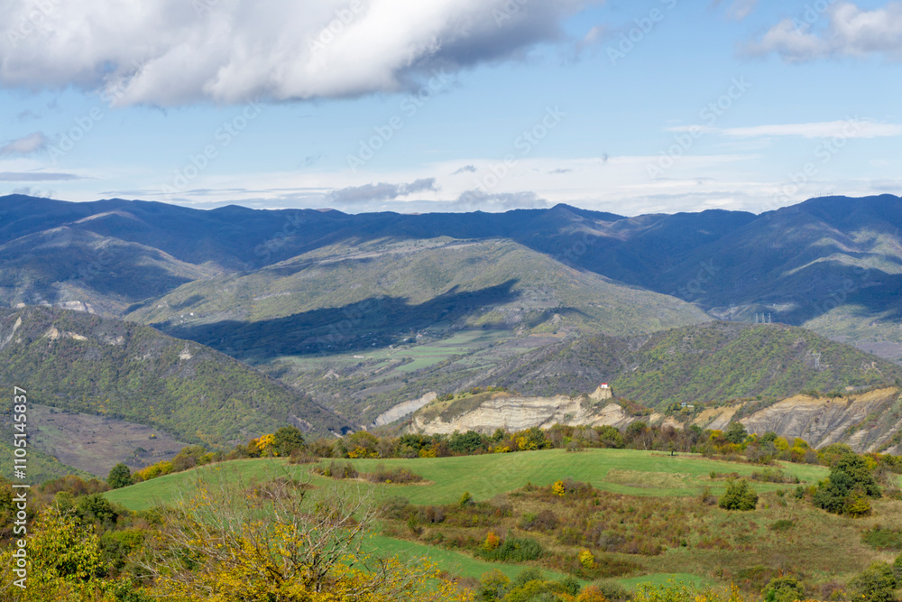 Fototapeta premium Green fields, autumn bushes illuminated by the sun. Mountains, hills and sky with clouds in the background