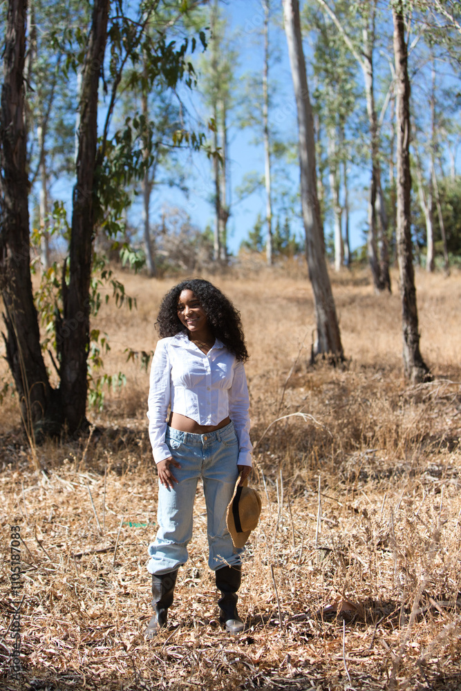 woman, young, beautiful, Latin, brunette with curly hair dressed in white shirt and jeans, boots and wearing a hat in her hand and walking among the eucalyptus trees in the countryside. Woman farmer. woman, young, beautiful, Latin, brunette with curly hair dressed in white shirt and jeans, boots and wearing a hat in her hand and walking among the eucalyptus trees in the countryside. Woman farmer.