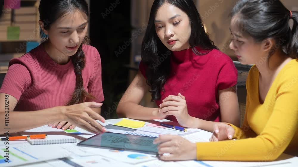 Three women are sitting at a table, working on a project. They are wearing red and yellow shirts Three women are sitting at a table, working on a project. They are wearing red and yellow shirts