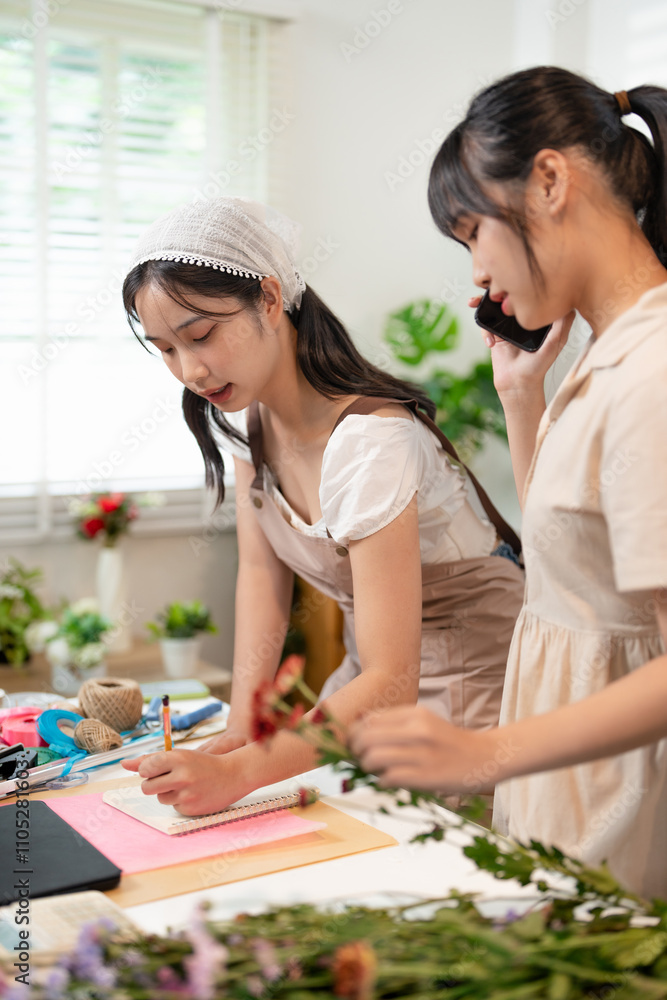 Female florist in apron checking details of order with colleague for creating and making flower bouquet with chrysanthemum in flower shop. Female florist in apron checking details of order with colleague for creating and making flower bouquet with chrysanthemum in flower shop.