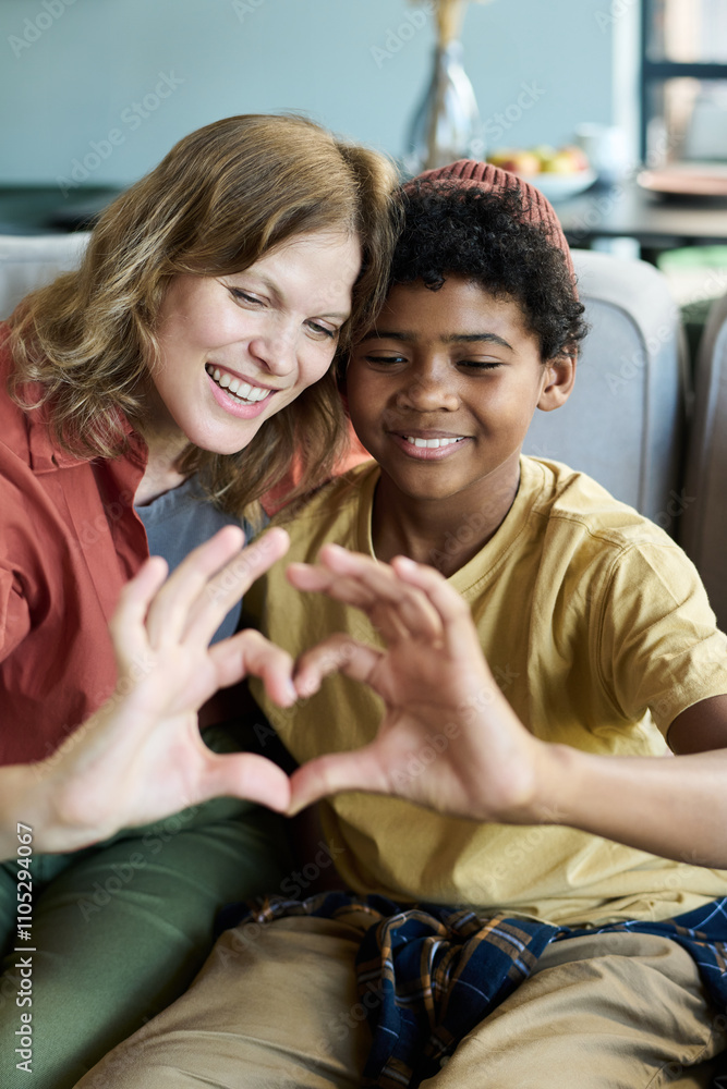 Mother and son forming heart shape gesture with hands while sitting on couch, sharing bonding moment and happiness. Caucasian mother and Black child smiling warmly Mother and son forming heart shape gesture with hands while sitting on couch, sharing bonding moment and happiness. Caucasian mother and Black child smiling warmly