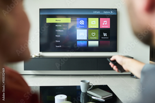 Naklejka Two people using remote control while seated and facing smart TV displaying various options on screen. Coffee table with mugs and remote control seen in foreground