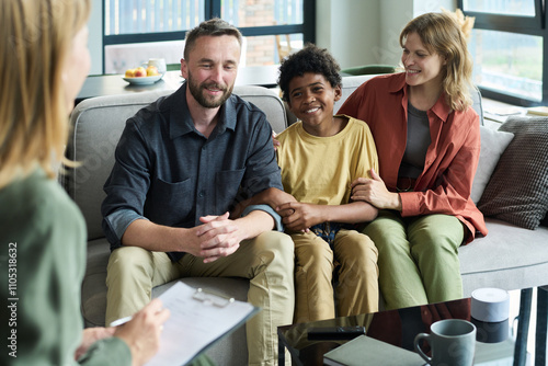 Multiracial family sitting on couch during counseling session, smiling and holding hands. Professional counselor in foreground with clipboard in hand and tablet on table