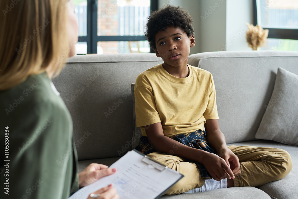 © pressmaster - Young child sitting on couch interacting with therapist holding clipboard engaging in conversation inside a cozy living room during therapy session © pressmaster - Young child sitting on couch interacting with therapist holding clipboard engaging in conversation inside a cozy living room during therapy session