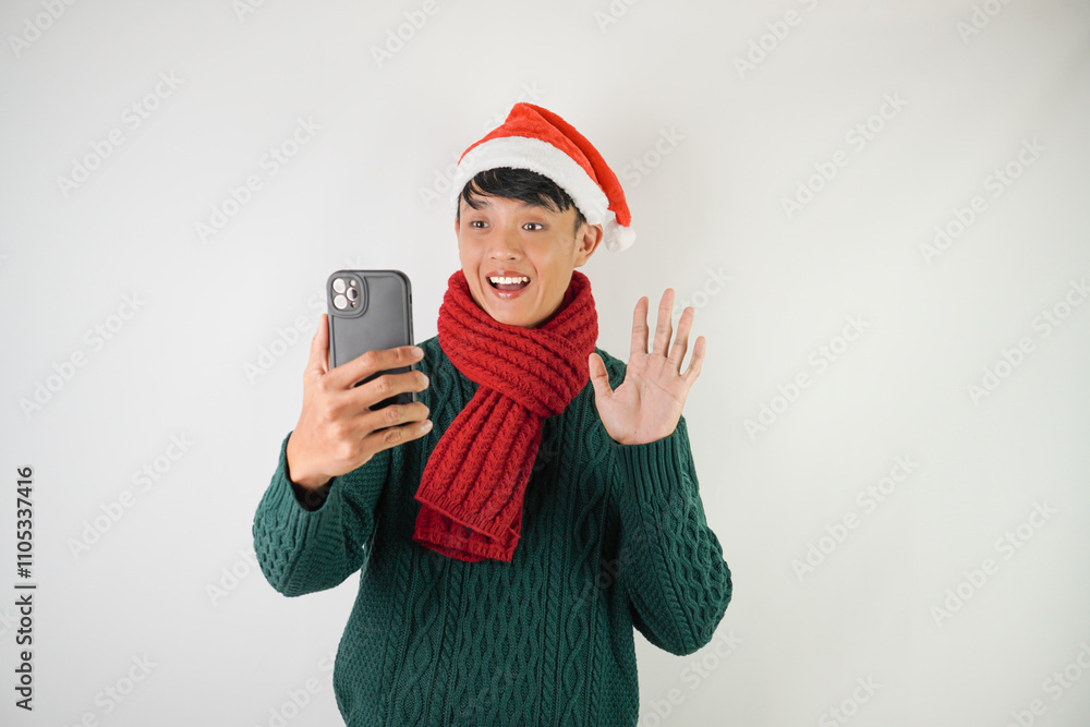 Young asian man wearing santa clause hat, red scarf and green long sleeved sweater is taking selfie using smartphone, isolated over white background. Young asian man wearing santa clause hat, red scarf and green long sleeved sweater is taking selfie using smartphone, isolated over white background.