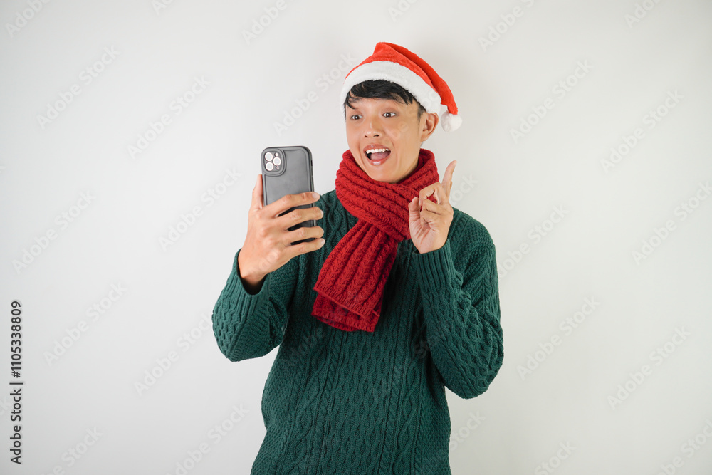 Young asian man wearing santa clause hat, red scarf and green long sleeved sweater is taking selfie using smartphone, isolated over white background. Young asian man wearing santa clause hat, red scarf and green long sleeved sweater is taking selfie using smartphone, isolated over white background.