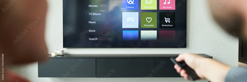 Two individuals seated on a couch are watching streaming media on a modern flat-screen TV mounted on wall. One person is holding remote control, interacting with device Two individuals seated on a couch are watching streaming media on a modern flat-screen TV mounted on wall. One person is holding remote control, interacting with device