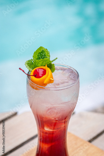 Singapore sling cocktail in glass garnished with Slice lemin and cherry and mint leaves on wooden planks in front of a pool photo