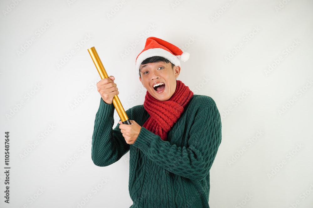 Young asian man wearing santa clause hat, red scarf and green long sleeved sweater with smiling face expression is holding confetti cannon, isolated over white background. Young asian man wearing santa clause hat, red scarf and green long sleeved sweater with smiling face expression is holding confetti cannon, isolated over white background.