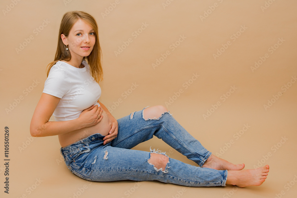 Young pregnant woman sitting on the floor in a white t-shirt and jeans, hugging her belly tenderly with her hands. Pregnancy concept, stylish motherhood. Copy space. Young pregnant woman sitting on the floor in a white t-shirt and jeans, hugging her belly tenderly with her hands. Pregnancy concept, stylish motherhood. Copy space.