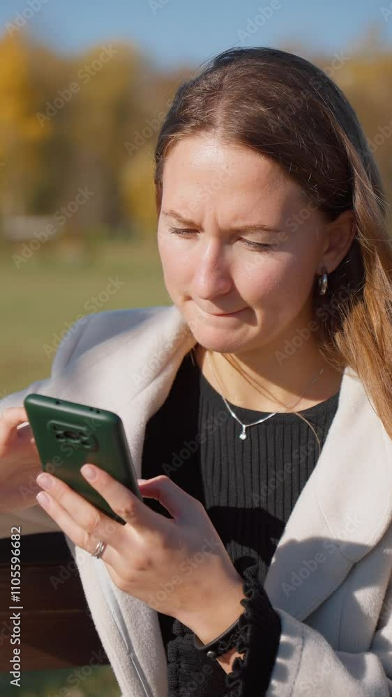 Young woman sitting on park bench reading text message on her smartphone, visibly angry as content is processed, reflecting emotions of frustration and irritation. Vertical video Young woman sitting on park bench reading text message on her smartphone, visibly angry as content is processed, reflecting emotions of frustration and irritation. Vertical video