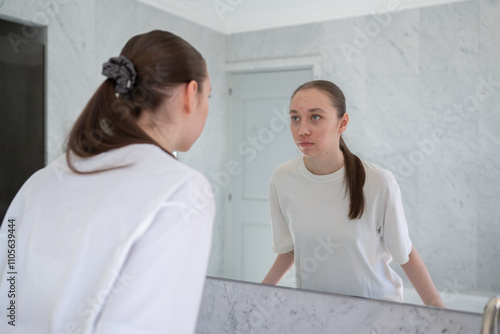 Gen z woman looking at her acne, stands before a mirror in bathroom. Concept of acne treatment, skincare, adult acne photo