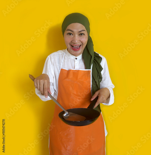 Young Asian Muslim Woman Housewife Wearing Kitchen Apron Cooking And Holding Pan And Spatula Isolated On Yellow Background