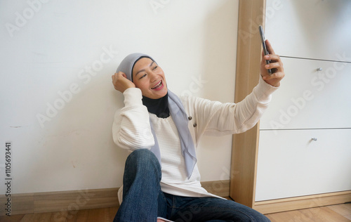 Asian Muslim Woman In A White Sweater And Hijab Is Sitting On The Floor And Taking A Selfie With Cell Phone.