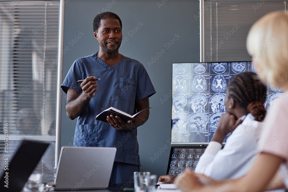 Fototapeta premium Professional male radiologist of African American ethnicity giving report on brain diagnostics with MRI scan to colleagues at board meeting, making notes in notebook at medical office
