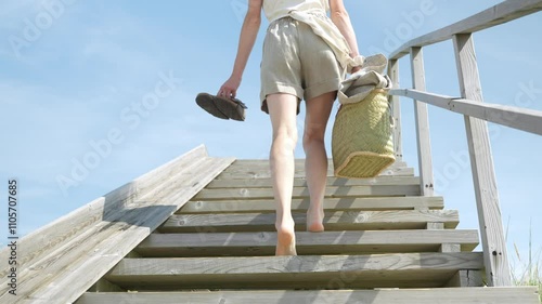 Slow motion camera view from the back, woman climbing up the beach stairs. Fit Caucasian female legs with linen shorts. Summer vacation mood, barefoot woman.