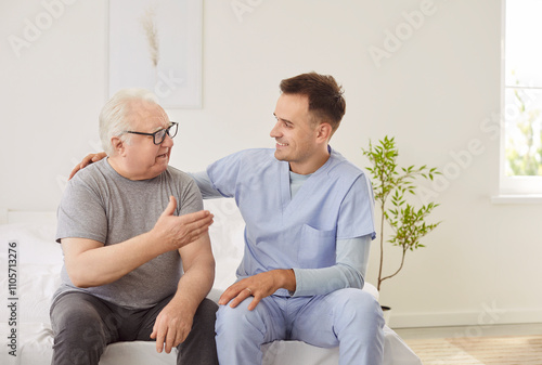 Friendly young male nurse talking with senior man after examining him in medical clinic. Elderly sick patient sitting on bed at hospital with caregiver. Nursing service and support concept.