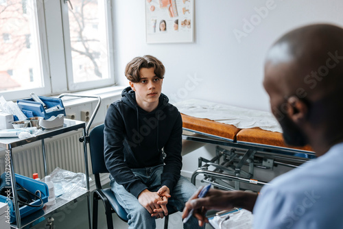 High angle view teenage boy consulting male medical expert while sitting on chair in examination room at clinic