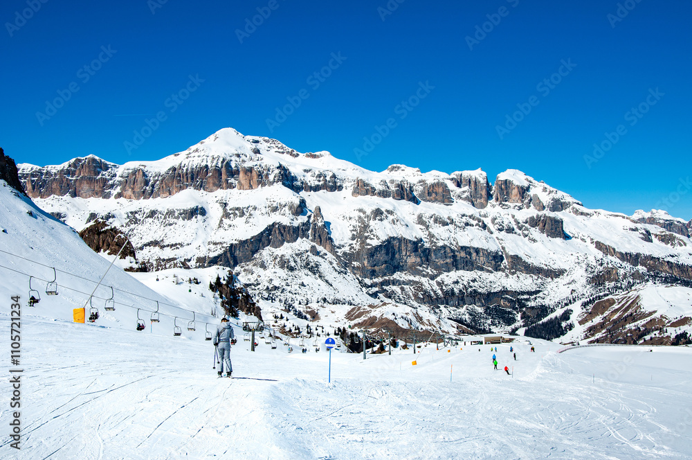 Easy ski slope with skiers and chairlift ski lift in Arabba ski resort in Dolomites, Italy. Sella Group mountain massif in the background. Blues sky with copy space