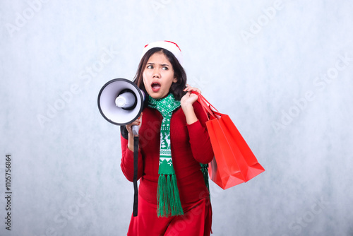 Charming woman wearing red christmas sweater, Santa hat and scarf, screaming angrily to the left hand holding megaphone loudspeaker and carrying red bag, isolated on white background photo