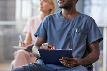 Cropped shot of African American man in blue scrubs keeping records of meeting on clipboard attending lecture in conference room while taking continuing learning course at hospital, copy space