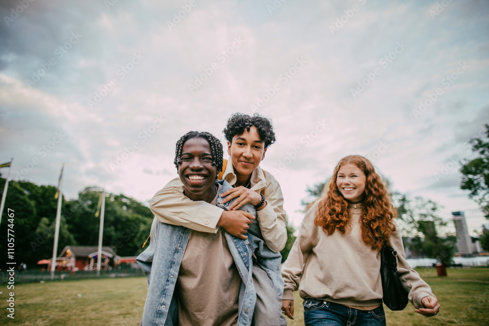 Teenage girl looking at braided hair boy piggybacking male friend at park Teenage girl looking at braided hair boy piggybacking male friend at park