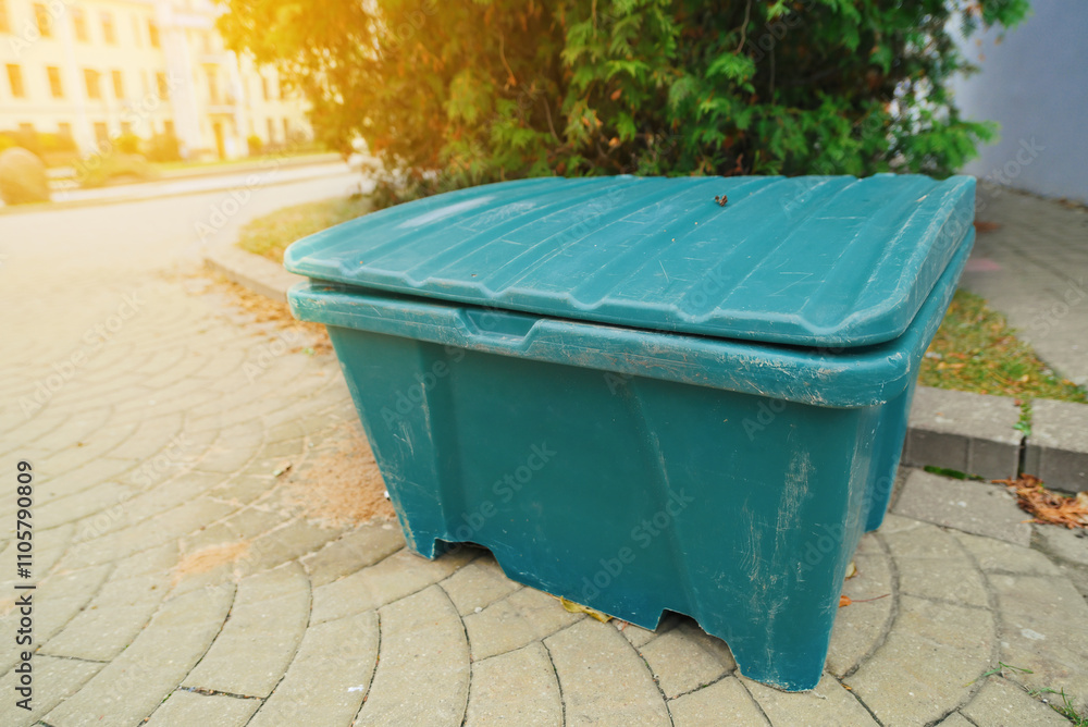 Green anti-slip sand storage container with an open lid placed on paved sidewalk, maintenance tools for pedestrian and road safety in winter. Utility sand box, used for street and sidewalk safety Green anti-slip sand storage container with an open lid placed on paved sidewalk, maintenance tools for pedestrian and road safety in winter. Utility sand box, used for street and sidewalk safety