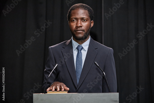 Portrait of adult African American male politician in formal wear holding hand on Constitution swearing allegiance to uphold law while standing at podium on stage against black curtains, copy space