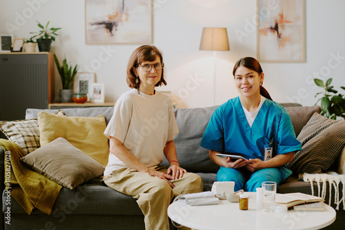 Portrait of smiling nurse in blue scrubs sitting on sofa with digital tablet, accompanied by elderly woman in white shirt, with cozy living room setting including various decor elements