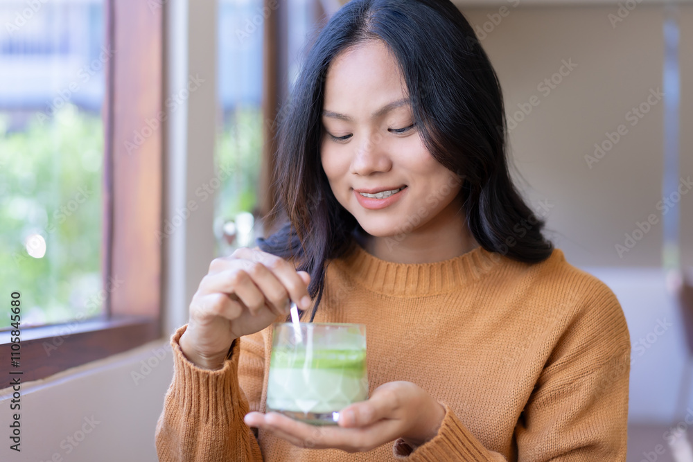 young woman smiling happy cheerful, and carefree Wear a sweater Sit by the window, hold a spoon, and stir the drink in the glass. Prepare to indulge favorite cup of green tea. Drink ideas. young woman smiling happy cheerful, and carefree Wear a sweater Sit by the window, hold a spoon, and stir the drink in the glass. Prepare to indulge favorite cup of green tea. Drink ideas.