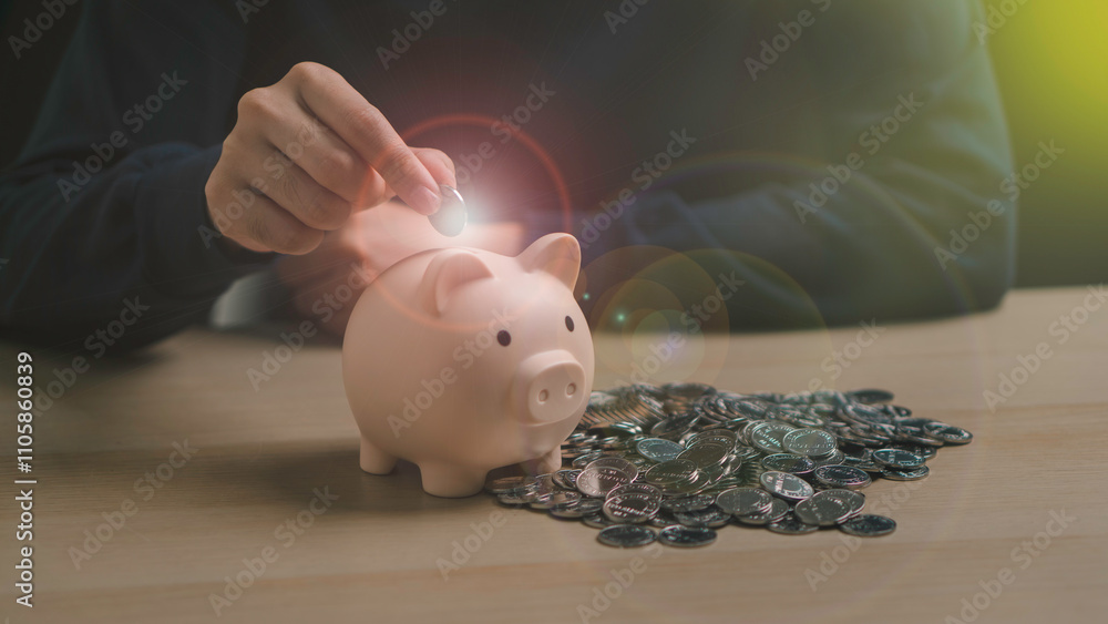A hand places a coin into a piggy bank surrounded by coins, representing saving money, long-term investment, and financial planning for the future.