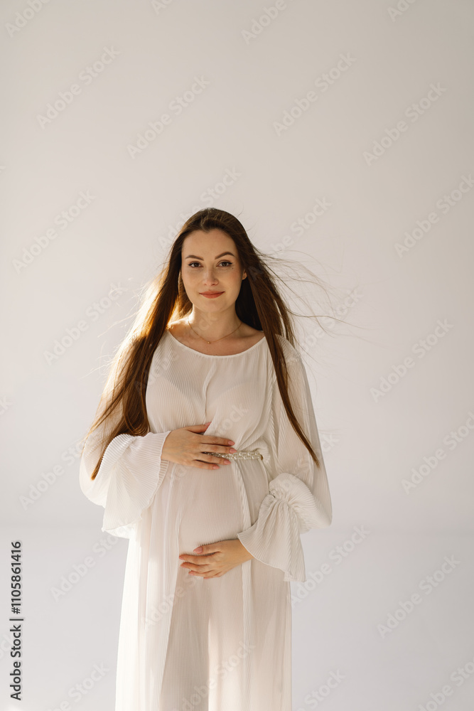 Cute young Pregnant girl sitting on the white studio in warm light with hands on pregnant stomach on wall background with shadow. Pregnant concept, free space, enjoying pregnancy. Pregnant body. Cute young Pregnant girl sitting on the white studio in warm light with hands on pregnant stomach on wall background with shadow. Pregnant concept, free space, enjoying pregnancy. Pregnant body.