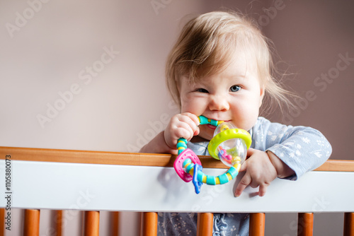 Cute Caucasian little baby girl standing in her crib and chewing on an infant toy, looking at the camera, with copy space. Funny baby