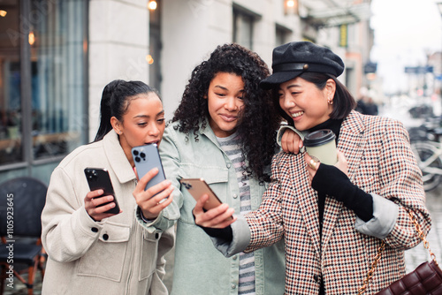 Three stylish women looking at phone and laughing together on city street photo