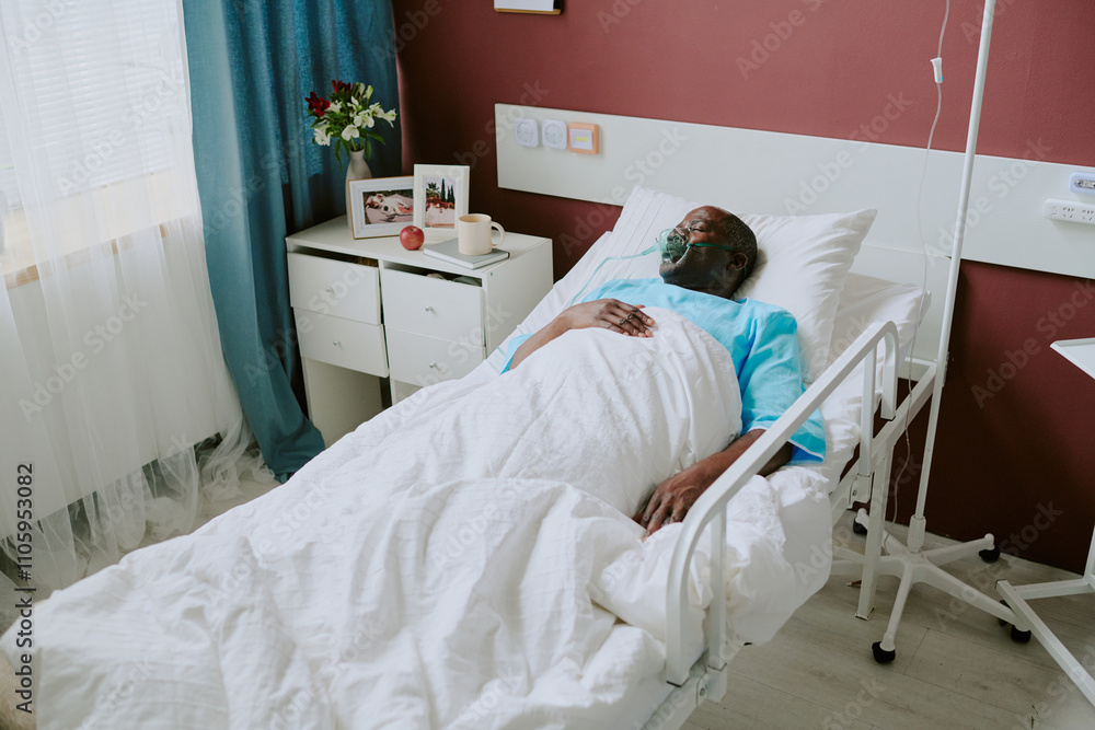 Hospital patient lying in bed beside window with light flowing in, surrounded by medical equipment and furnishings, wearing face mask for breathing assistance Hospital patient lying in bed beside window with light flowing in, surrounded by medical equipment and furnishings, wearing face mask for breathing assistance
