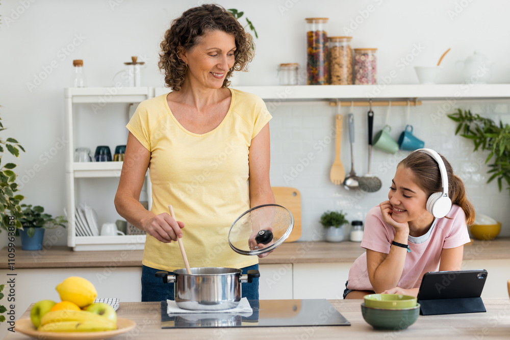 Mature cheerful mother cooking some soup while talking with her kind daughter in the kitchen Mature cheerful mother cooking some soup while talking with her kind daughter in the kitchen