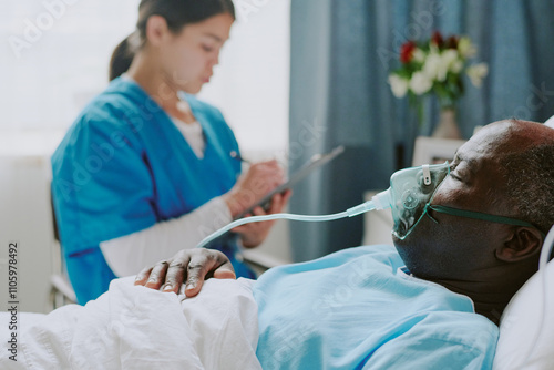 African American patient resting in hospital bed while being attended by Latin nurse writing observations on clipboard in brightly lit room with flowers in background