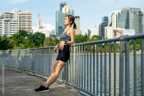 Side view of Asian woman lean wall railing and look at her watch to relax after exercise in public garden with high building of big city in background and soft light.