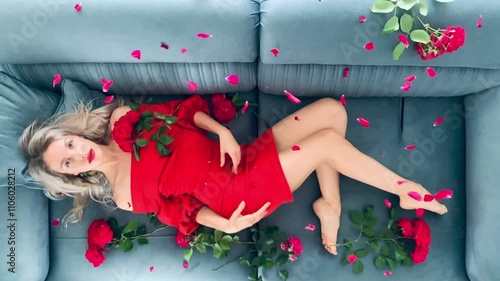 Beautiful sexy hot blonde woman in a red dress, surrounded by tea rose flowers, lies on the bed. Red rose petals fall on the hot beauty from above. Slow motion.