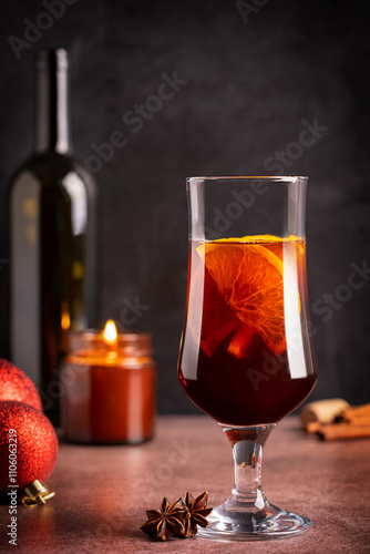 One glass of traditional homemade winter hot red mulled wine drink or spiced grog cocktail made with orange slices, cinnamon and anise served on table with christmas baubles, bottle and candle light
