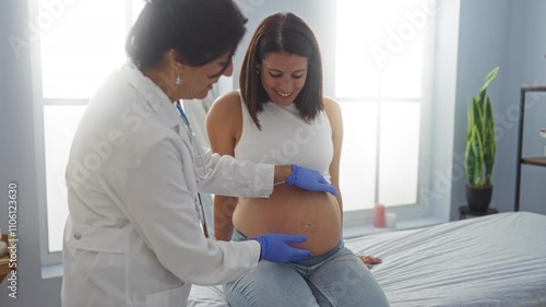 Doctor examining pregnant woman at clinic with woman smiling and touching belly in indoor medical setting with sunlight in background photo