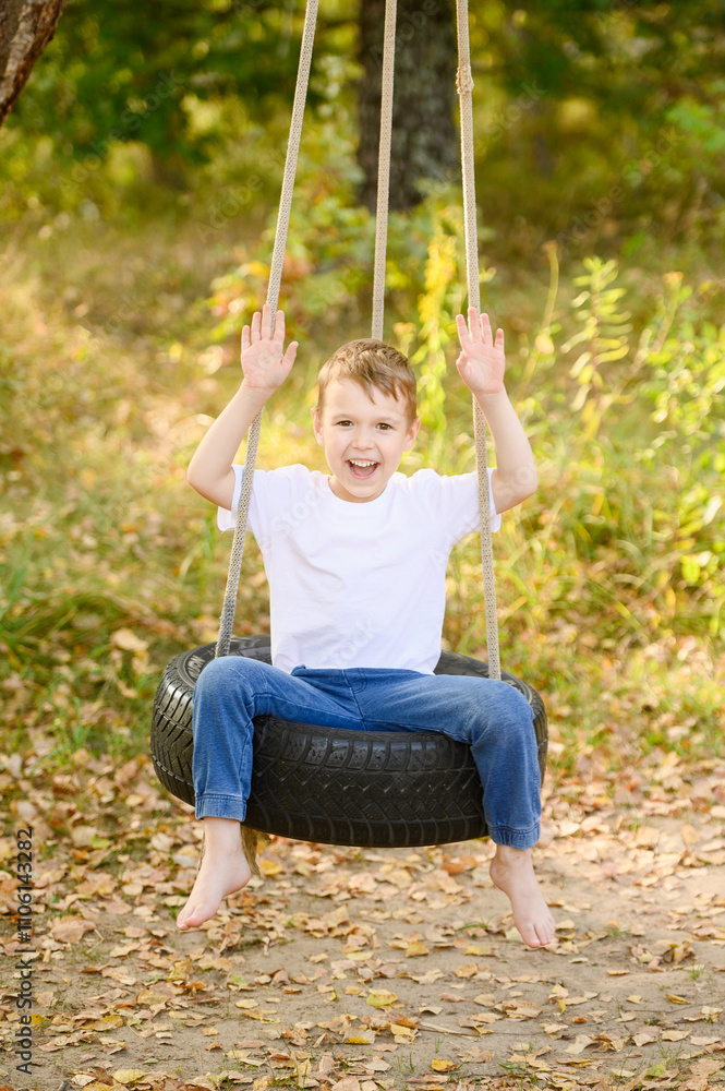 Boy swings on swing, sitting on wheel suspended from tree, smiling happily and raising arms. Happy boy playing outside, enjoying country vacation.Child swings on rubber tire from car wheel.Game safety Boy swings on swing, sitting on wheel suspended from tree, smiling happily and raising arms. Happy boy playing outside, enjoying country vacation.Child swings on rubber tire from car wheel.Game safety