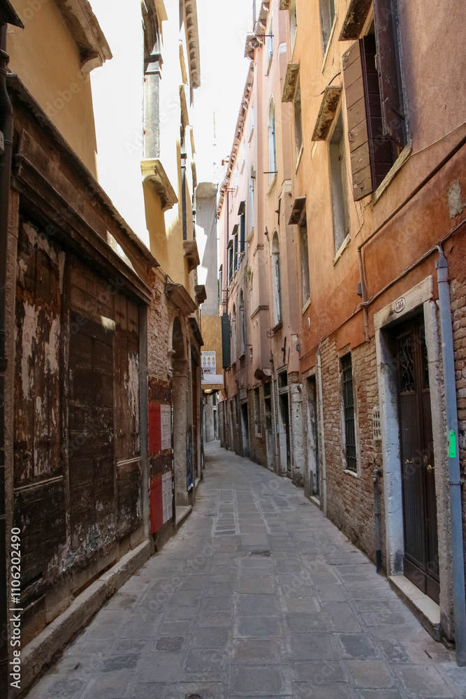 Fototapeta premium Side view of empty narrow pedestrian street between residential brick buildings in a sunny summer day in Venice, Italy. Soft focus. Copy space. Old town Italian architecture. Vacation in Europe theme.