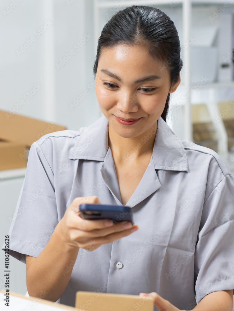 Asian female small business shop owner using application on smartphone scanning QR code on parcel box for checking shipment information and order number to prepare for delivery at warehouse office. Asian female small business shop owner using application on smartphone scanning QR code on parcel box for checking shipment information and order number to prepare for delivery at warehouse office.