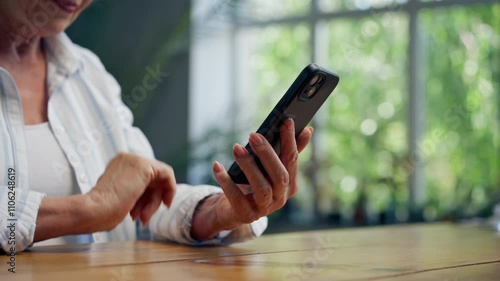 hands close-up of a very elderly gray-haired grandmother in a light blouse with a slight smile sitting against the background of a blue wall with large indoor plants writing a message on the phone